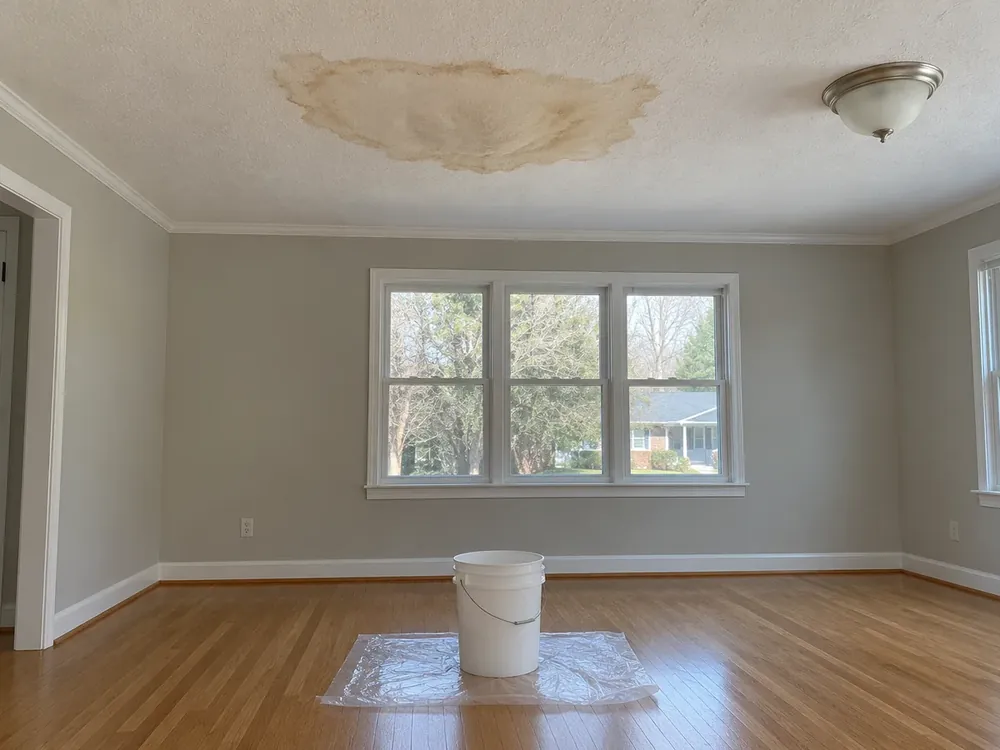 Brown water stain on a living room ceiling with a bucket and towel on the floor beneath